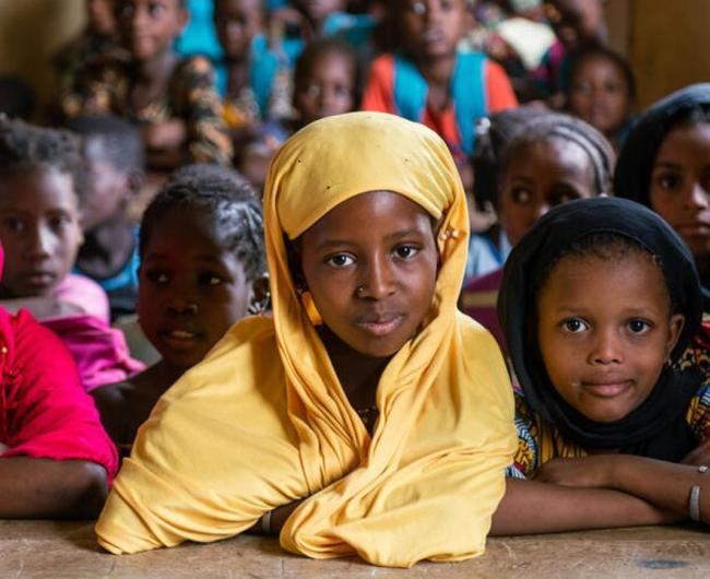 A group of girls in hijabs sit together and look to camera. A girl in the center of the photo looks straight to camera in a brilliant yellow hijab.