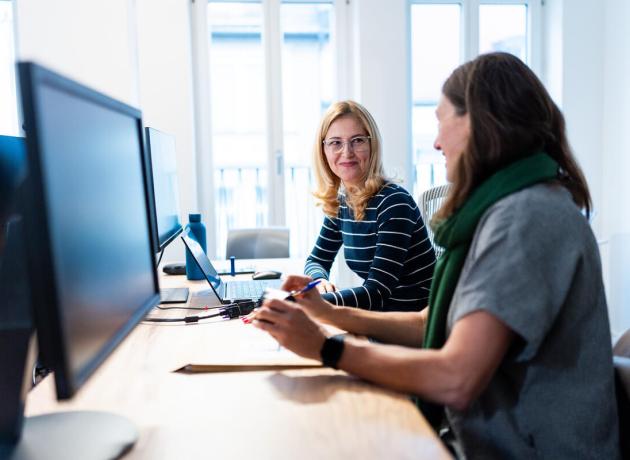 Two women sitting side by side at a long desk in front of computers, looking at each other in conversation. 